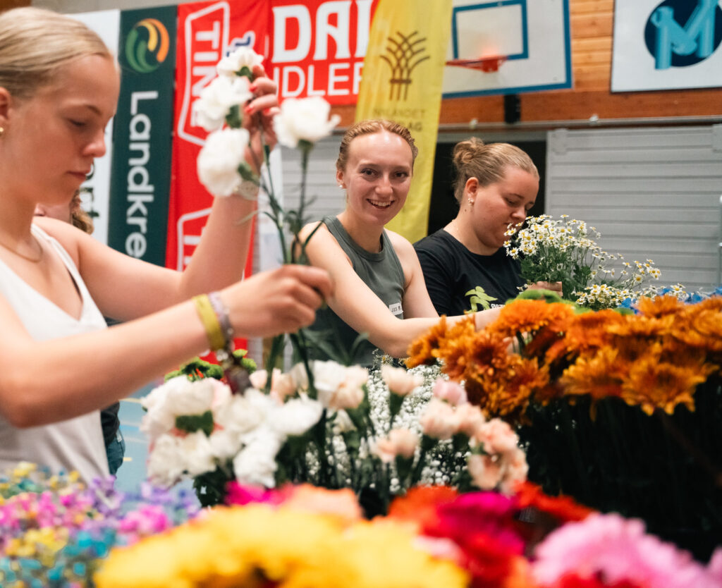 Bilde av tre tevlere som tevler i NM i blomsterdekorering. De plukker blomster i en idrettshall. Den ene tevleren ser i kamera.