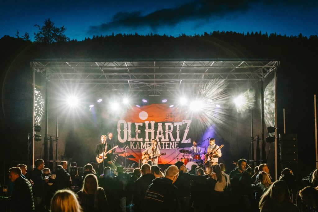 Bilde av OLE HARTZ og kameratene på scenen under en konsert. Det er mange i publikum. Det er mørkt ute.