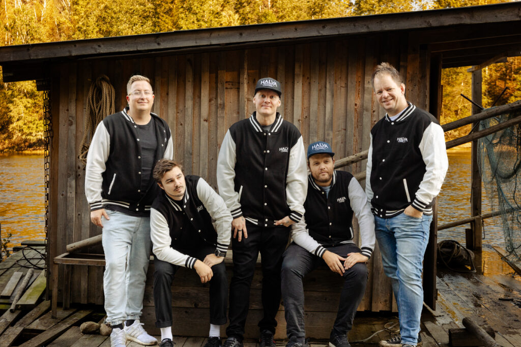 Bandmedlemmene står på rekke foran et båthus i matchende jakker. Noen smiler og ser i kamera mens noen smiler og ser i luta. to av de sitter.